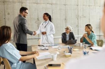 Two people shake hands, a physician and person dressed in business attire while other people watch intrigued